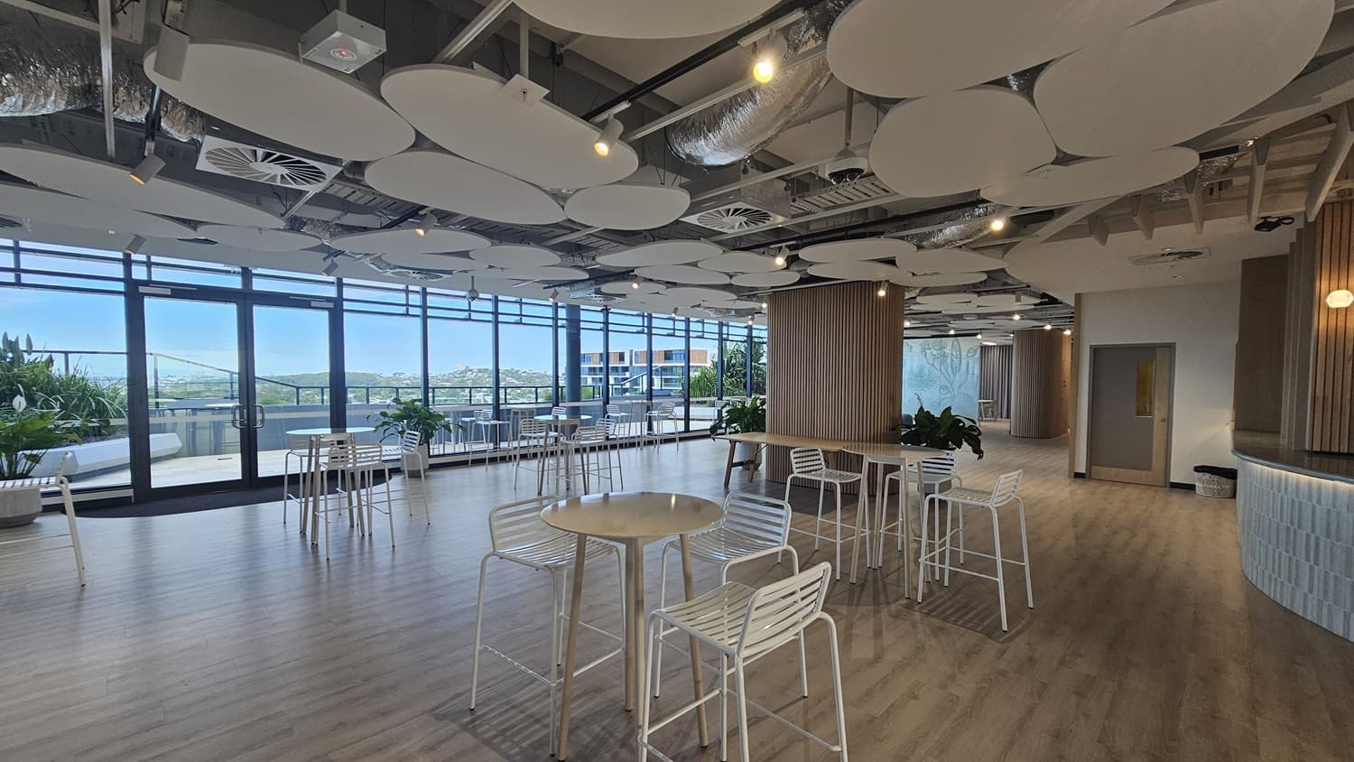 An open space with a large window on the left and a small bar on the right with a tiled counter. The space is furnished with round tables and white chairs. The ceiling has wood panels and many large, white, circular panels. The floor is wood laminate.