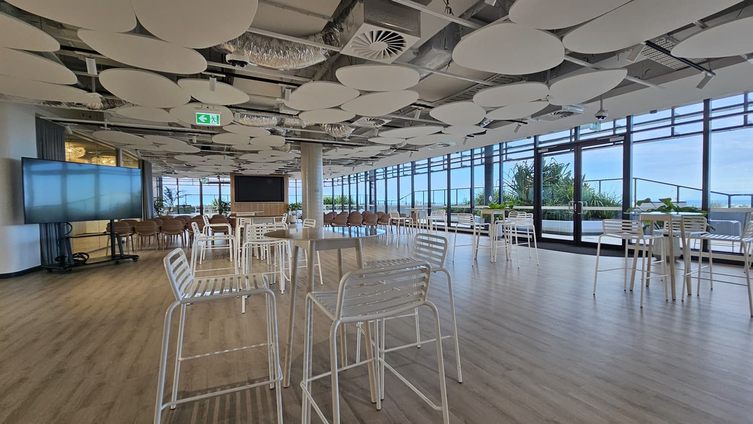 An open space with large windows on the right looking out over a city with water in the distance. There are many white tables and chairs throughout the space, and a large TV screen on the left. The ceiling has a unique design with many large, white, circular panels. The floor is wood laminate.