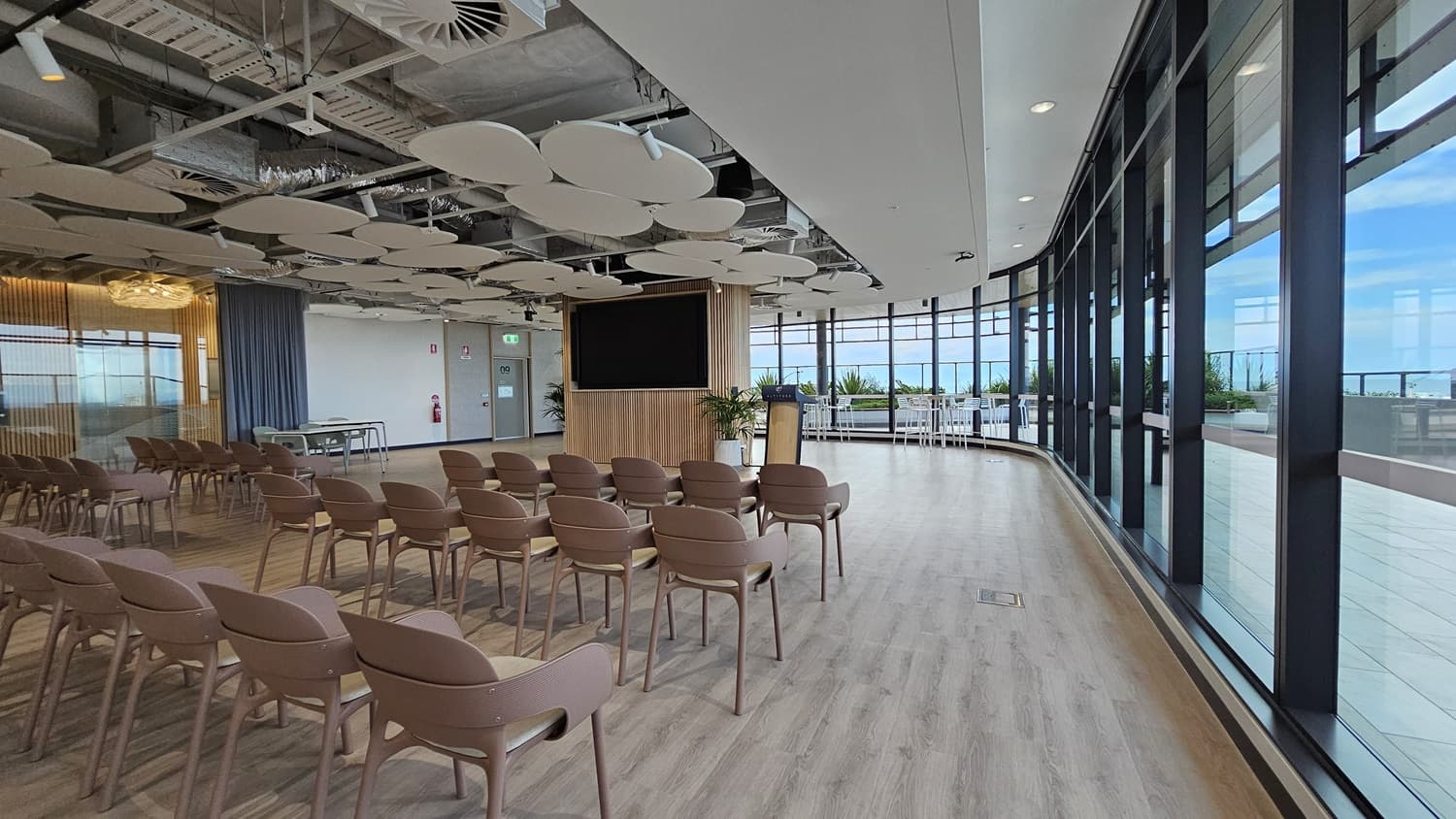 An empty conference room with rows of brown chairs facing a large TV screen. There is a wooden panel behind the screen and a small wooden podium with a microphone. The room has a large window on the right, looking out over a balcony and a city. The ceiling has many large, white, circular panels. The floor is wood laminate.