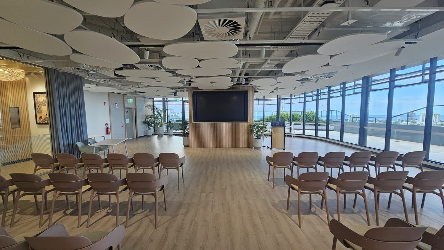 A large, empty room with rows of brown chairs facing a large TV screen. The screen is in front of a wooden panelled wall. There is a small wooden podium with a microphone. On the right are large windows with a view of a balcony and a city. The ceiling has many large, white, circular panels. The floor is wood laminate.