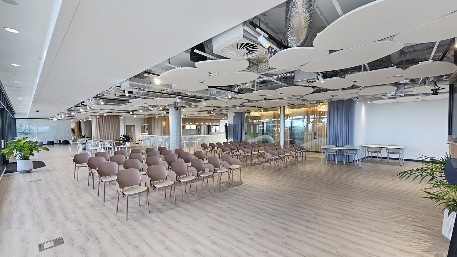 An empty room with rows of brown chairs facing a large, empty space. On the right is a large window overlooking a city. In the centre of the room there are two tables with chairs, and a large potted plant in the corner of the room. The ceiling has white, circular panels. The floor is wood laminate.