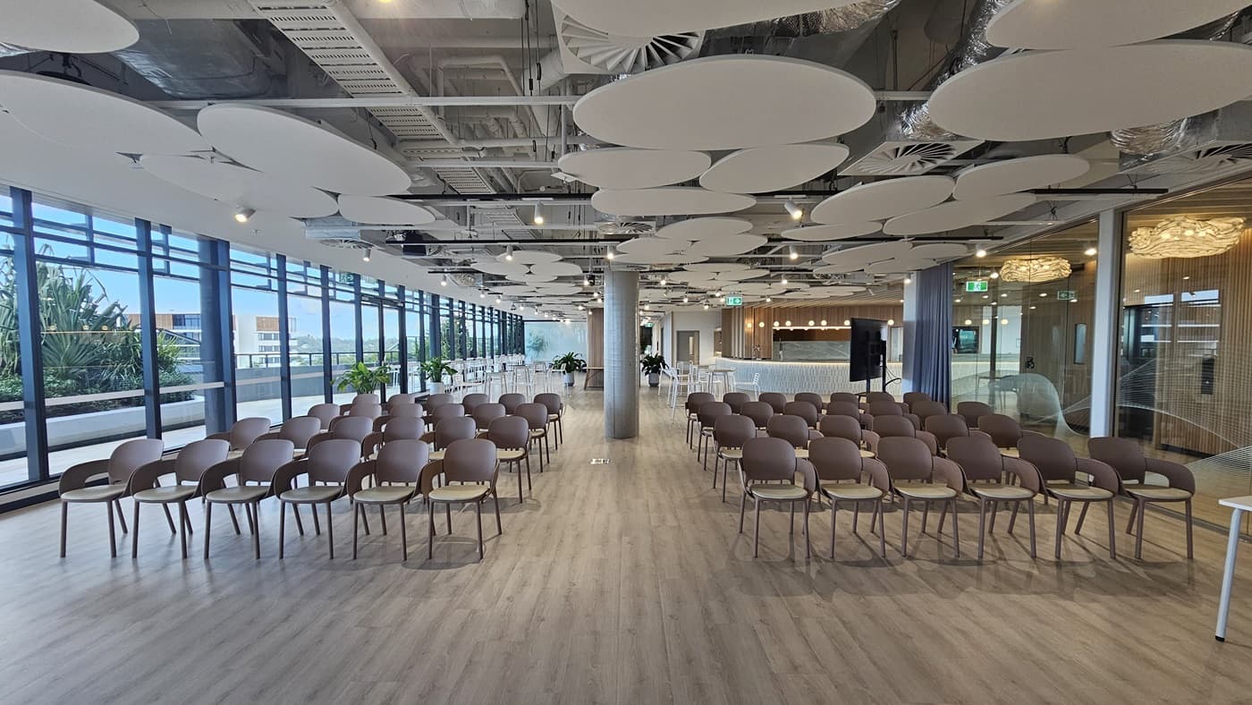 A large room with rows of brown chairs facing each other. On the left are large windows with a view of a balcony and a city. There is a pillar in the middle of the room. The ceiling has many large, white, circular panels. The floor is wood laminate.