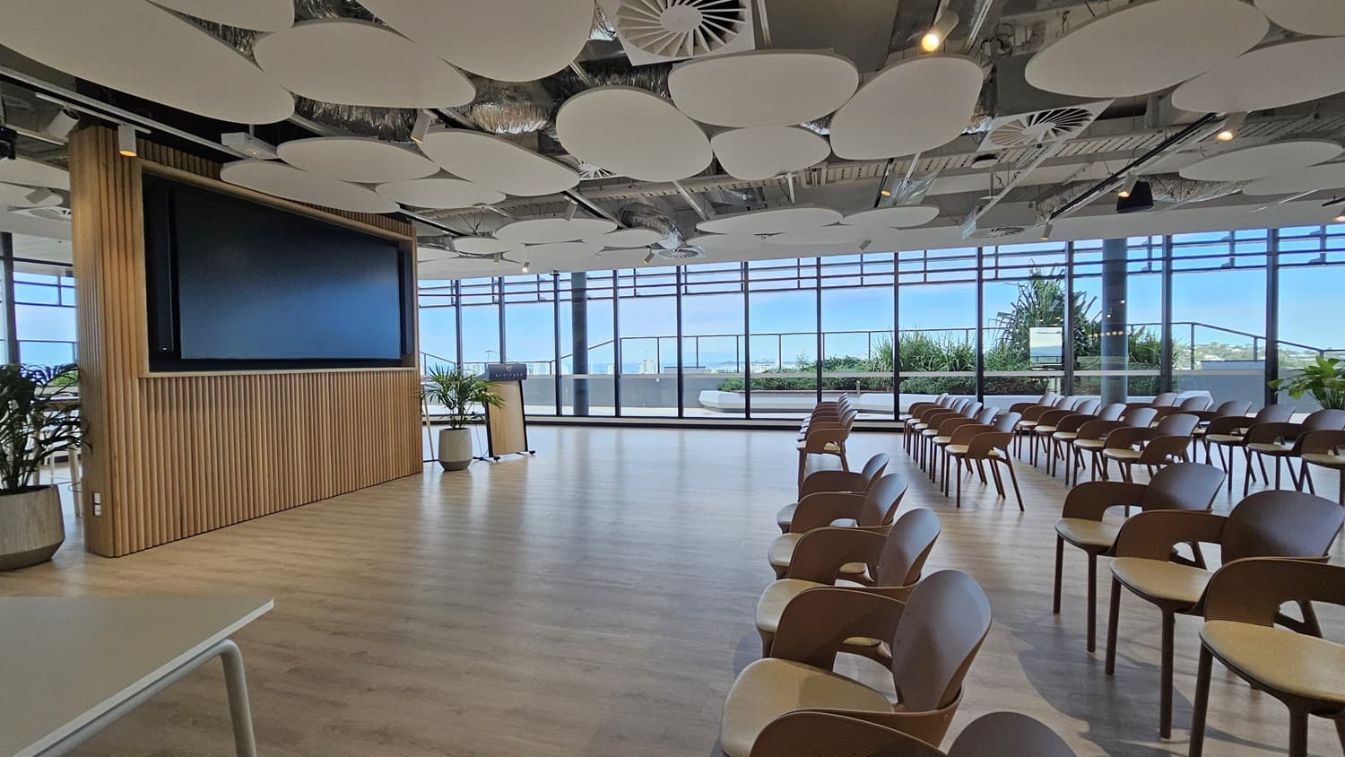 A large room with a large TV screen on the left wall. The wall is made of wood slats. There are rows of brown chairs facing the screen. There are large windows on the right, overlooking a city. The ceiling is white with many large, round panels. The floor is wooden.