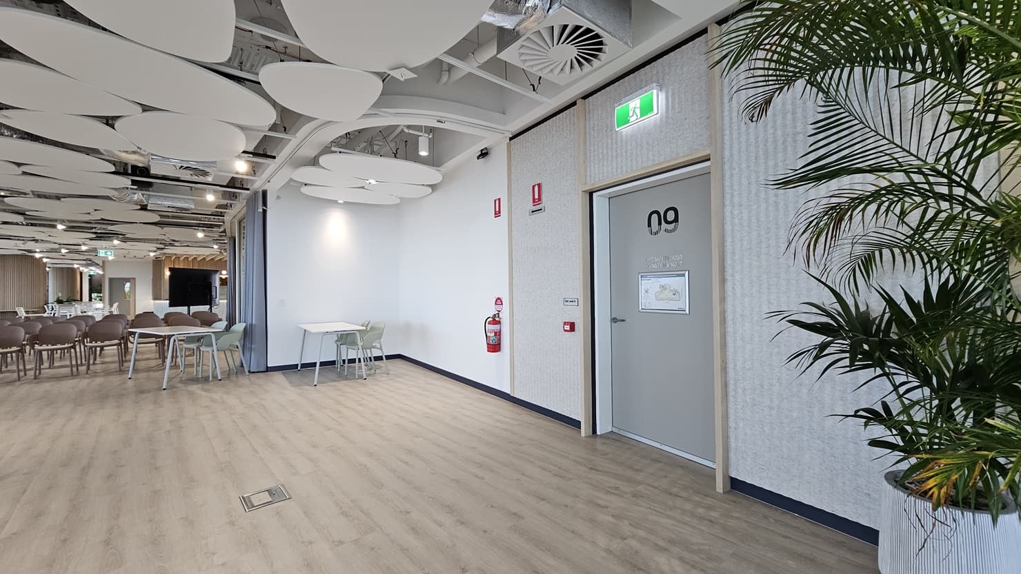 A hallway with a large, open space to the left. The space has rows of chairs and tables. On the right is a wall with a door labelled '09' and a large plant in a white pot. The ceiling has many large, white, circular panels. The floor is wood laminate.