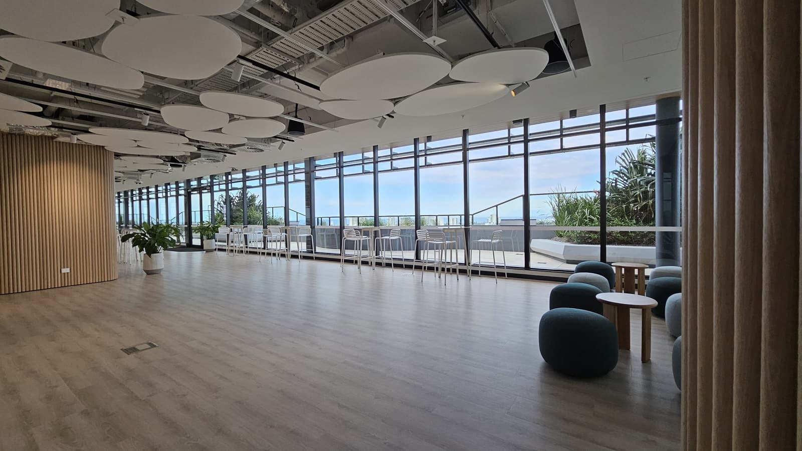 A large, open room with large windows on the left looking out onto a balcony. In the centre of the room are some small, grey pouffes and a glass wall with a botanical design. To the right is a hallway with a door labelled '09'. The ceiling has many large, white, circular panels. The floor is wood laminate.
