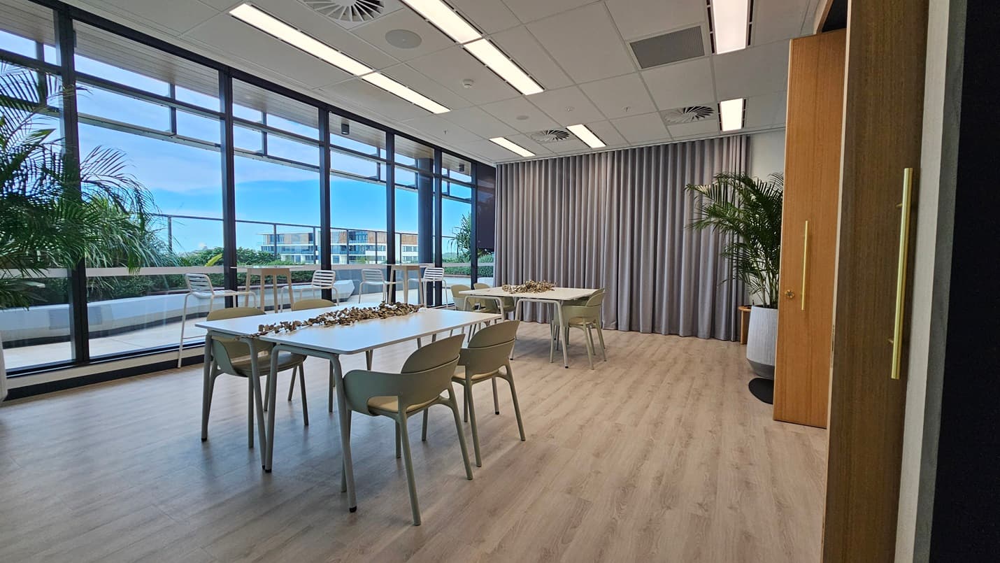 A meeting room with two white tables and chairs in the centre. On the left are large windows with a view of a balcony and a city. To the right of the room is a grey curtain and a wooden door. There is a large plant in a white pot next to the door. The ceiling has white rectangular light panels. The floor is wood laminate.
