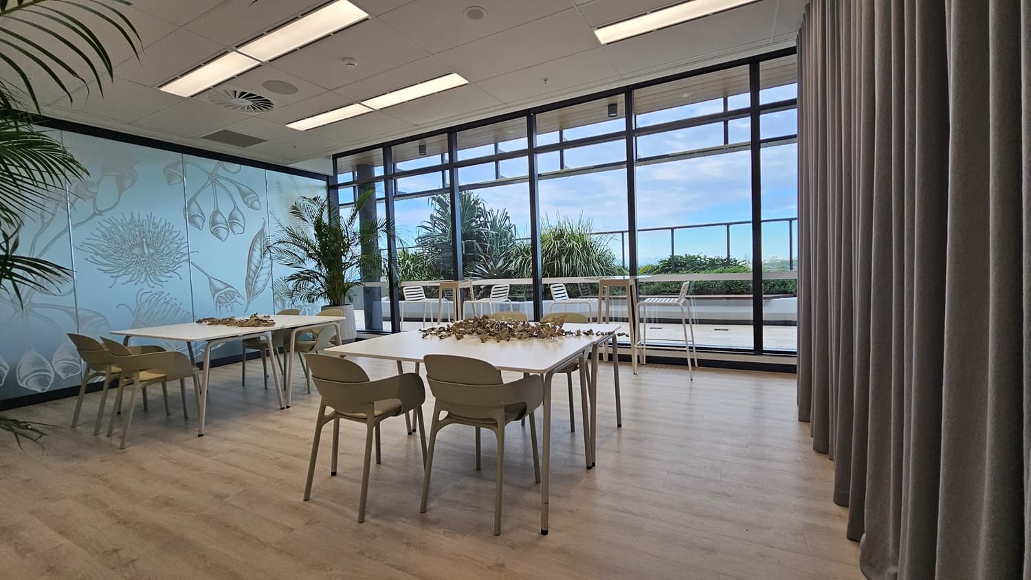 A meeting room with two white tables and chairs in the centre. On the left is a wall with a frosted glass panel featuring a floral design and a green plant in a pot. On the right, large windows offer a view of a balcony and a cityscape beyond. Grey curtains hang to the right of the windows. The ceiling has white rectangular light panels. The floor is wood laminate.