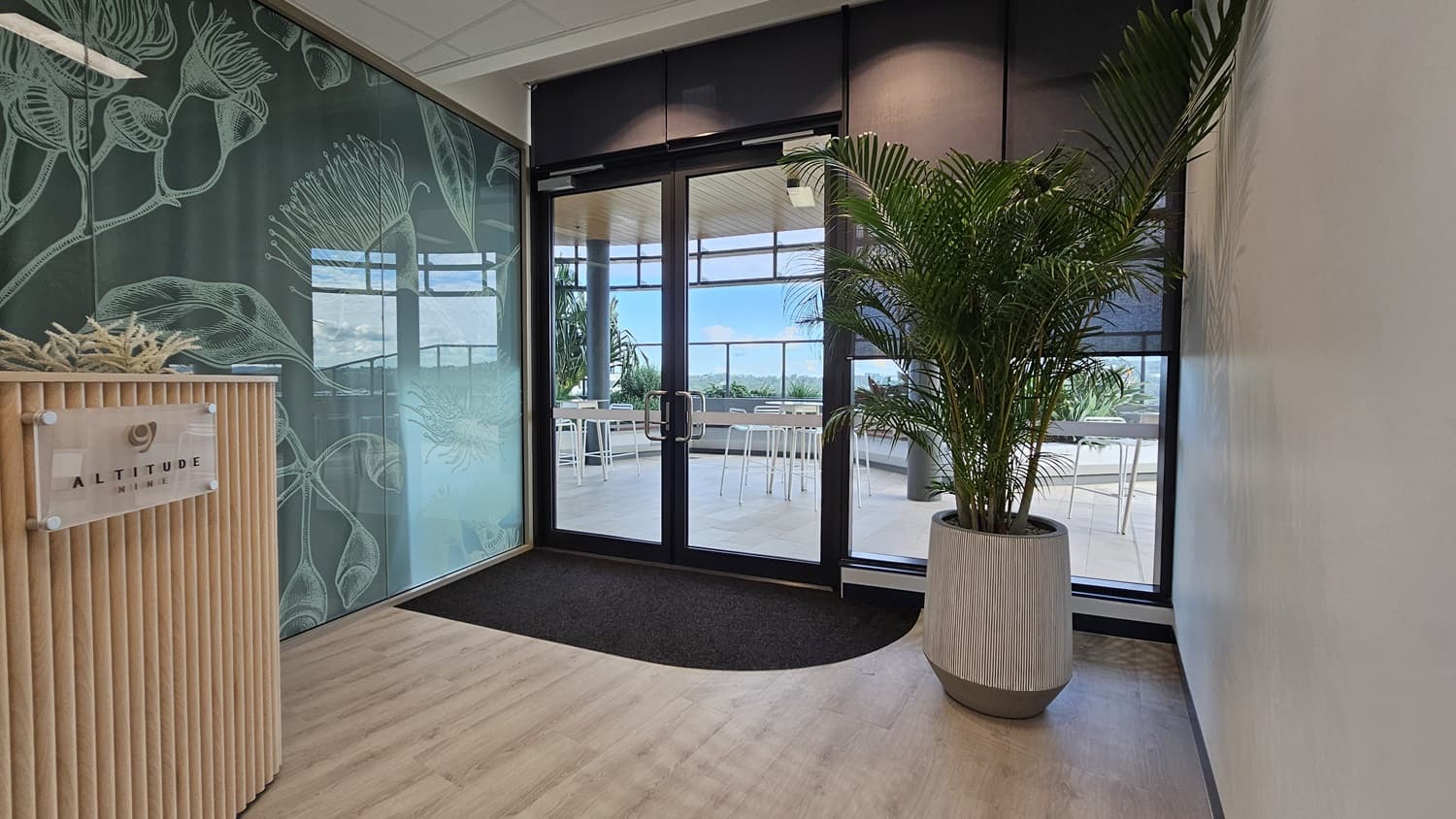 A reception area with a wooden reception desk on the left with a sign that reads "Altitude Nine". The wall behind the desk has a frosted glass panel with a botanical design. On the right are double doors leading to a balcony with a view of a city. There is a large plant in a white pot to the right of the doors. The floor is wood laminate.