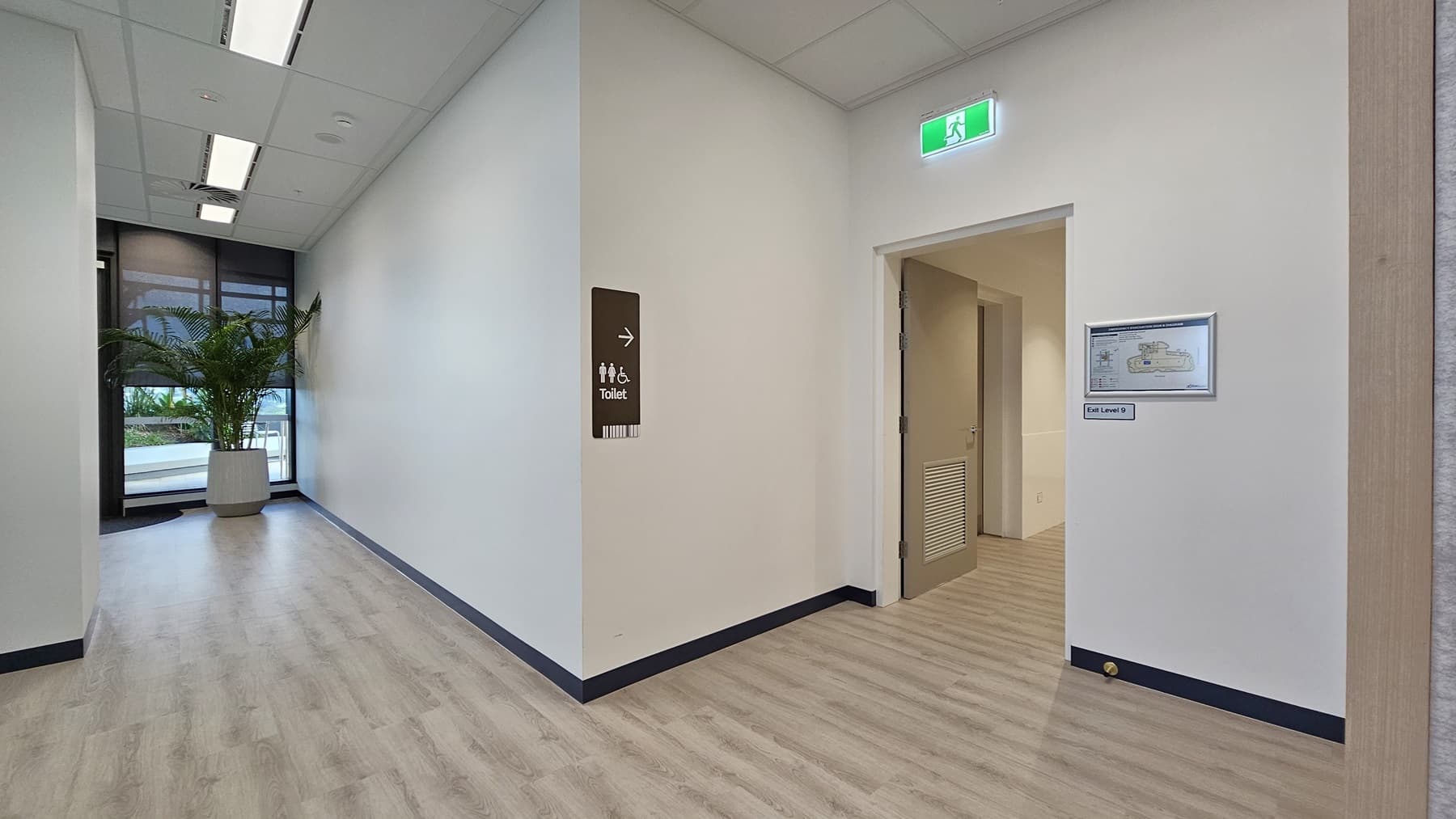 A narrow hallway with a plant in a pot on the left. There is a window behind the plant. On the right, there is a door labelled 'Toilet' and a doorway with a green emergency light above it. The ceiling has white light panels. The floor is wood laminate.