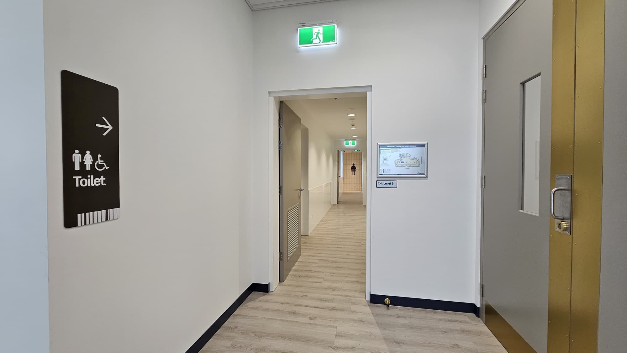 A hallway with a sign on the left that says 'Toilet' with an arrow pointing right and symbols for male, female, and disabled access. There is a door on the left and a doorway leading to another hallway. On the right is a large grey door with a brass strip. A green emergency light is above the doorway to the right. The floor is wood laminate.