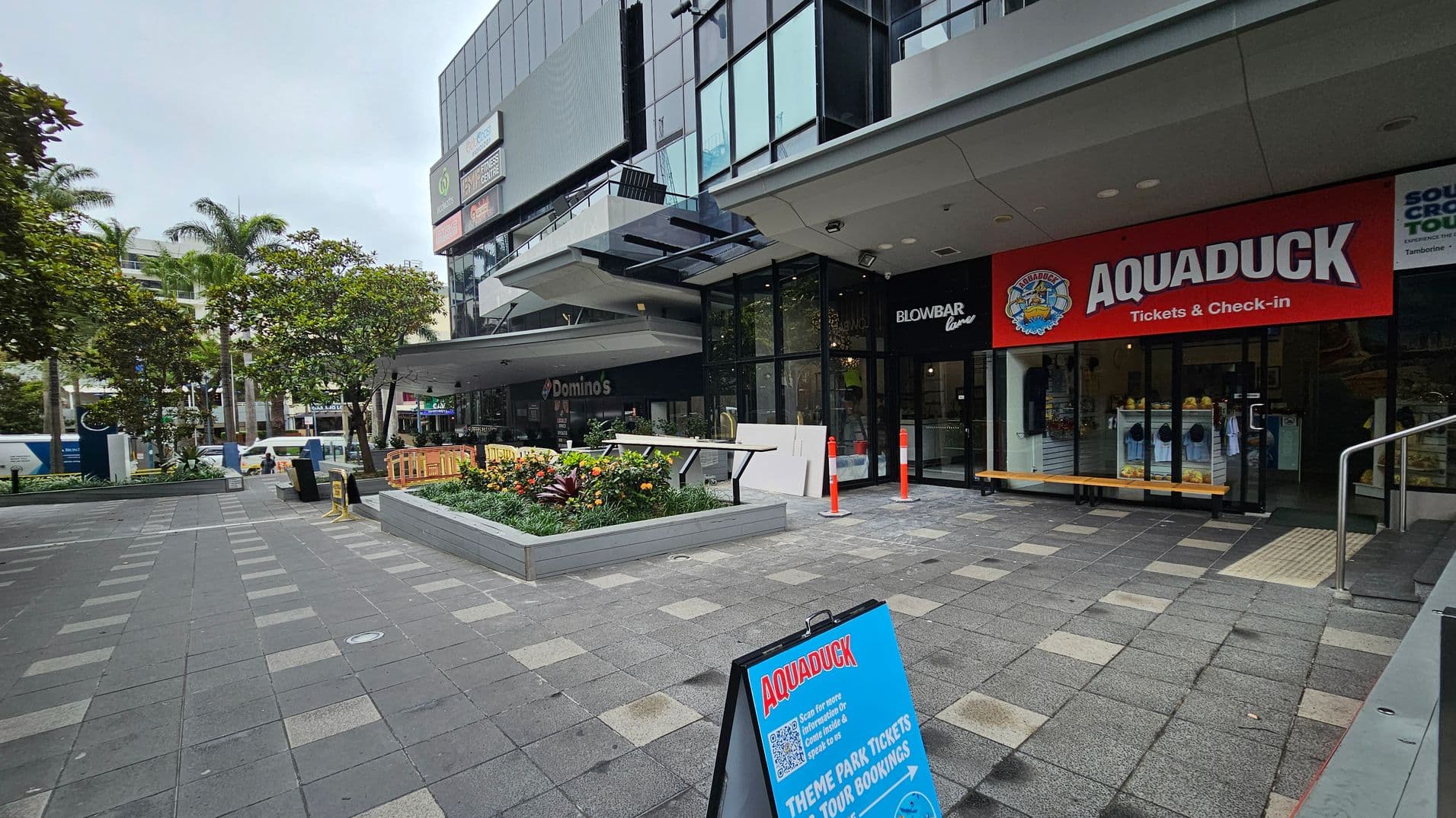 On the right, there's a building with a red sign for "Aquaduck Tickets & Check-in" and a glass entrance. A blue sandwich board sign is on the pavement in front. To the left, there's a Domino's sign and a Blowbar Lane sign above. In the middle, a raised planter with flowers and small trees is surrounded by a tiled pedestrian area. In the background, more buildings and palm trees are visible. The pavement is grey with lighter square patterns.