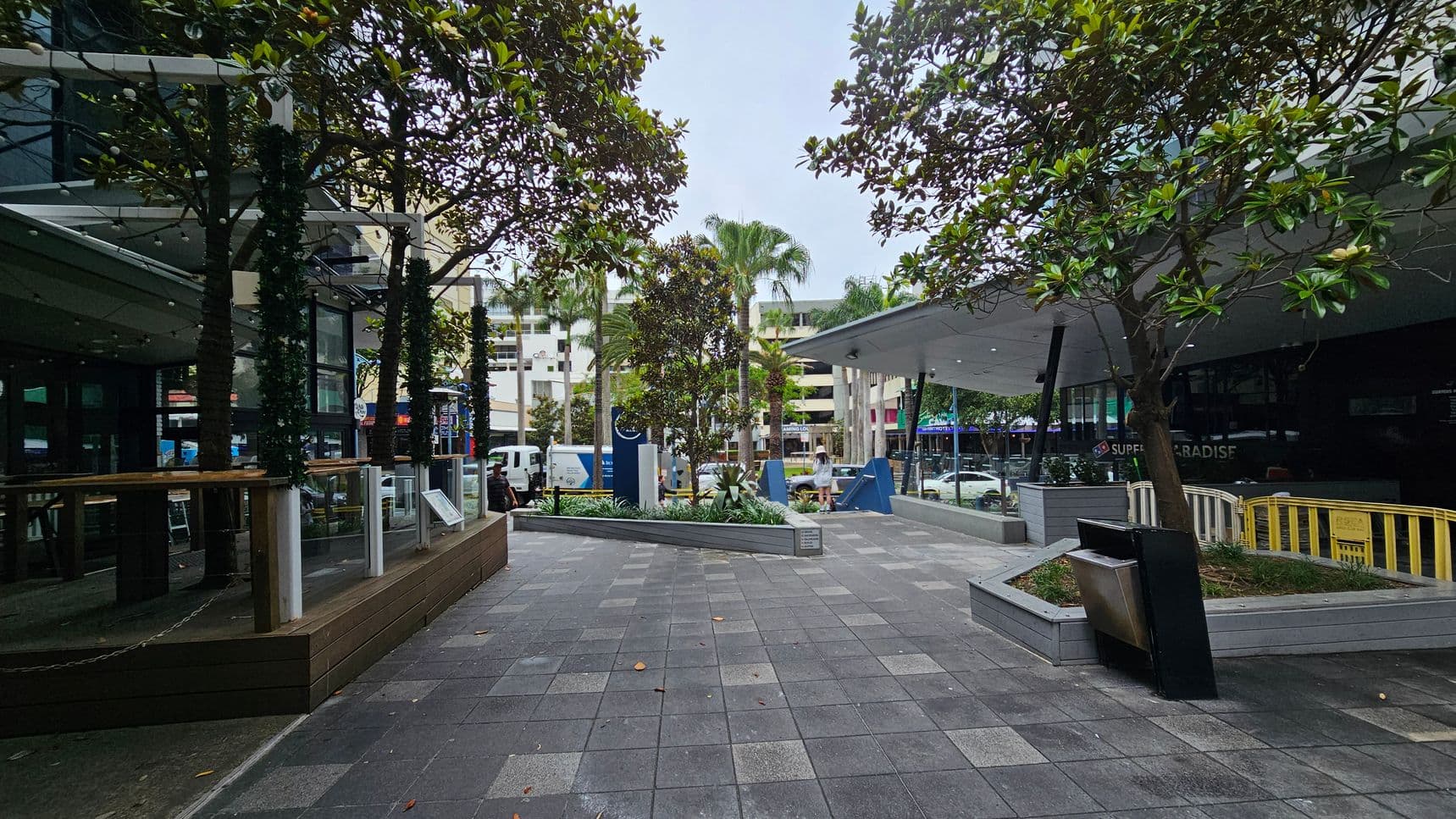 A paved urban area with trees and planters on both sides. On the left, there is a building with glass windows and a covered outdoor seating area. On the right, a modern structure with a slanted roof and more seating. In the background, a street with parked vehicles and a multi-storey building. The ground is covered with square tiles, and there are decorative lights wrapped around the trees.