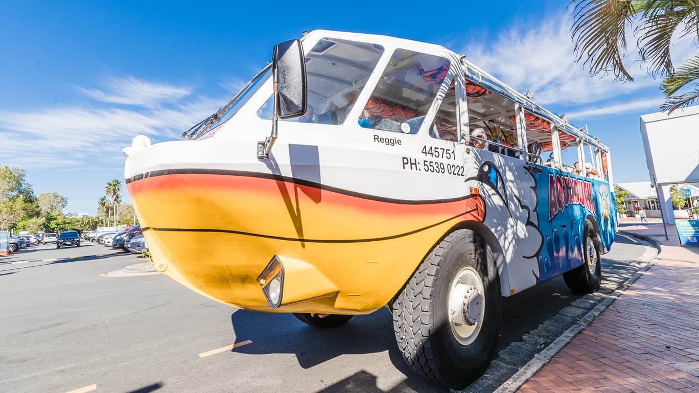 Side of the Aquaduck parked near a red brick pathway that is partly shaded by palm trees. Detailed below the right glass window of the driver’s corner is a black typeface that reads “Reggie 445751” and “PH: 5539-0222”. On the ceiling inside the vessel are orange life jackets. The side body has an orange-white color scheme, blue waves, a logo, and the word “AQUADUCK.” Left of the Aquaduck is the car park that has an alternating black and yellow speed bump. To the back are two white buildings.