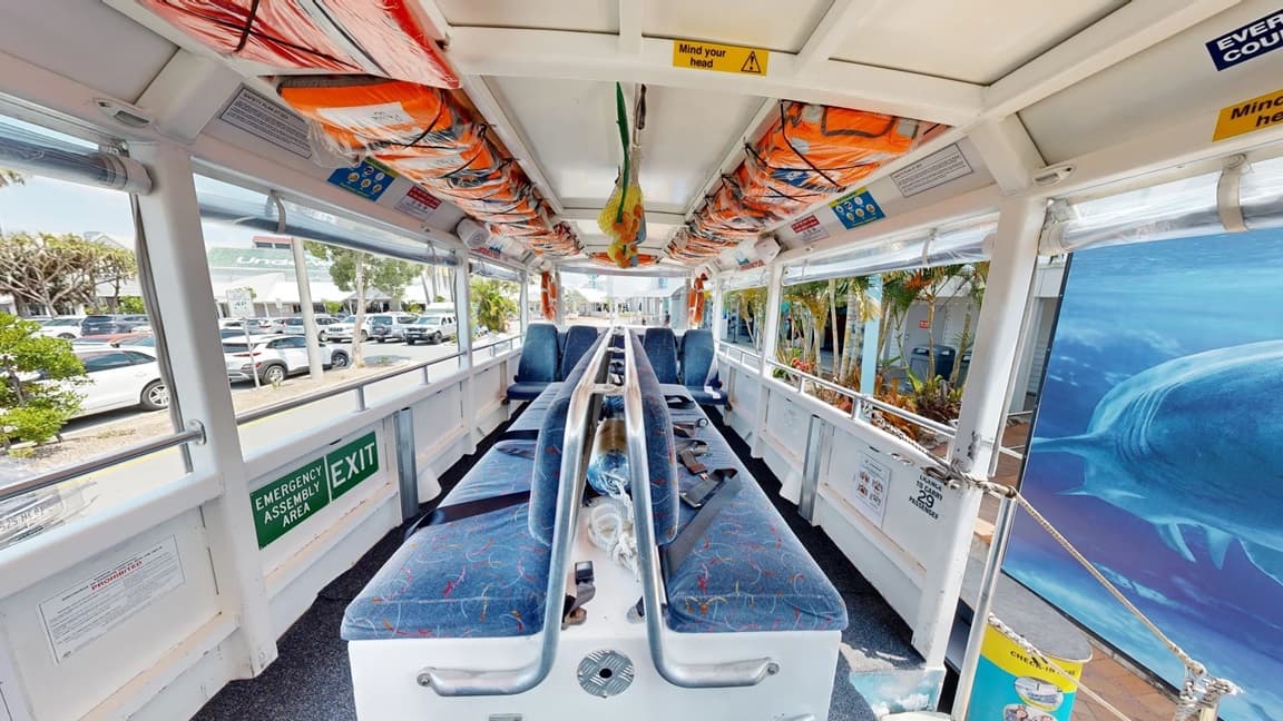 The middle of the Aquaduck is a long bench-type seat with a back support that has blue seat padding with yellow, red, and light blue line patterns. To the back is another row of 4 blue seats with yellow, red, and light blue line patterns, which are separated by a center aisle. Above and below the windows are varying colors and sizes of signs for rules and safety precautions. On the ceiling are two lanes of orange life vests inside plastic secured by black straps forming an X.