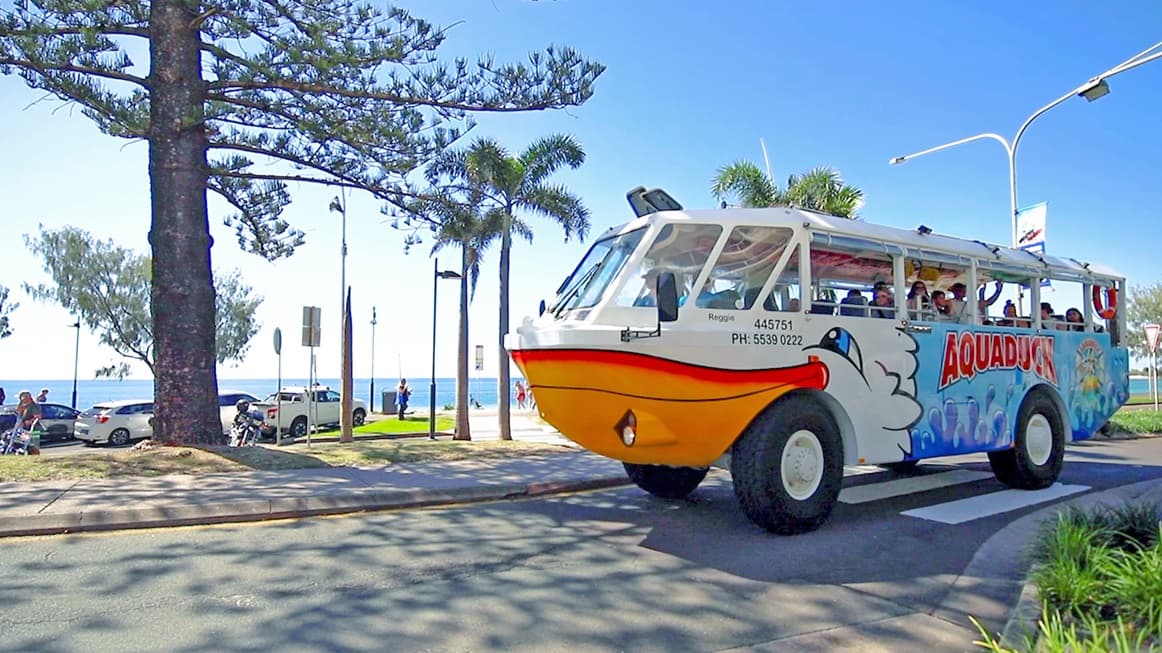 Aquaduck passing through the white zebra crossing in SEA LIFE Sunshine Coast. The vessel has orange-white color schemes, blue waves, a logo, and the word “AQUADUCK” on the side. The front resembles the head of a smiling duck with a big yellow beak. On the right is a verge garden with grasses that have parallel venation. To the left is a large pine tree and behind it is a car park, a pathway with palm trees on its edge, and parking signs, with the ocean in the background.