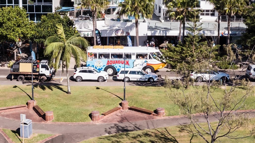 Arial view of the road in front of a white building with a glass handrail terrace on the second floor and a park nearby. The focal point is the large white bus labeled “Aquaduck” in red lettering over a light blue background occupying the left lane while the right has traffic of white automobiles and a truck. On top of the bus are three light brown rectangular platforms. Next to the busy street is a park with a brown V bench pattern with circular ends that have vertical lines.