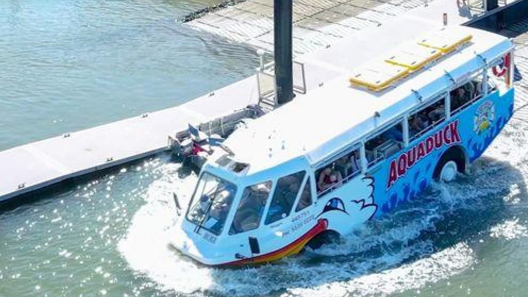 Aquaduck descending down a steep boat ramp. A visible difference in the floor from grey to cement color can be seen in the image. The background features a clear blue sky, a body of water, buildings, vehicles, and a palm tree on the right. The vessel has orange-white color schemes, blue waves, a logo, and the word “AQUADUCK” on the side. The front resembles the head of a smiling duck with a big yellow beak. Found on the end of the vessel are two orange life buoys.