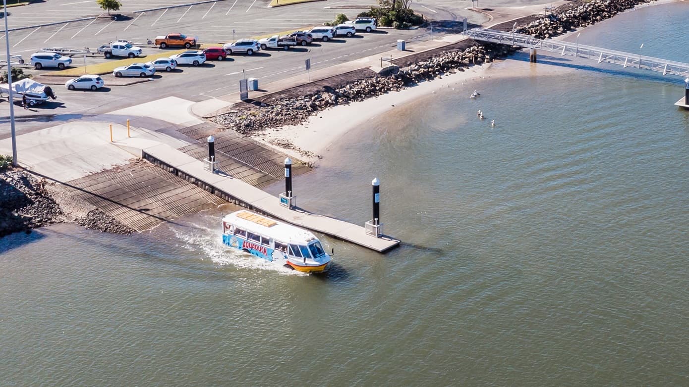 Aerial view from the left side of the Aquaduck leaving the steep boat ramp with horizontal lines. In the middle of the ramp is a smooth bridge that has a short ramp entry access with two yellow bollards. The bridge ends at a deeper portion of the Mooloolah River. On the right side of the bridge are three black posts with a white cone top. Further to the back of the ramp is a parking lot where several cars are parked adjacent to the rocky shorelines.