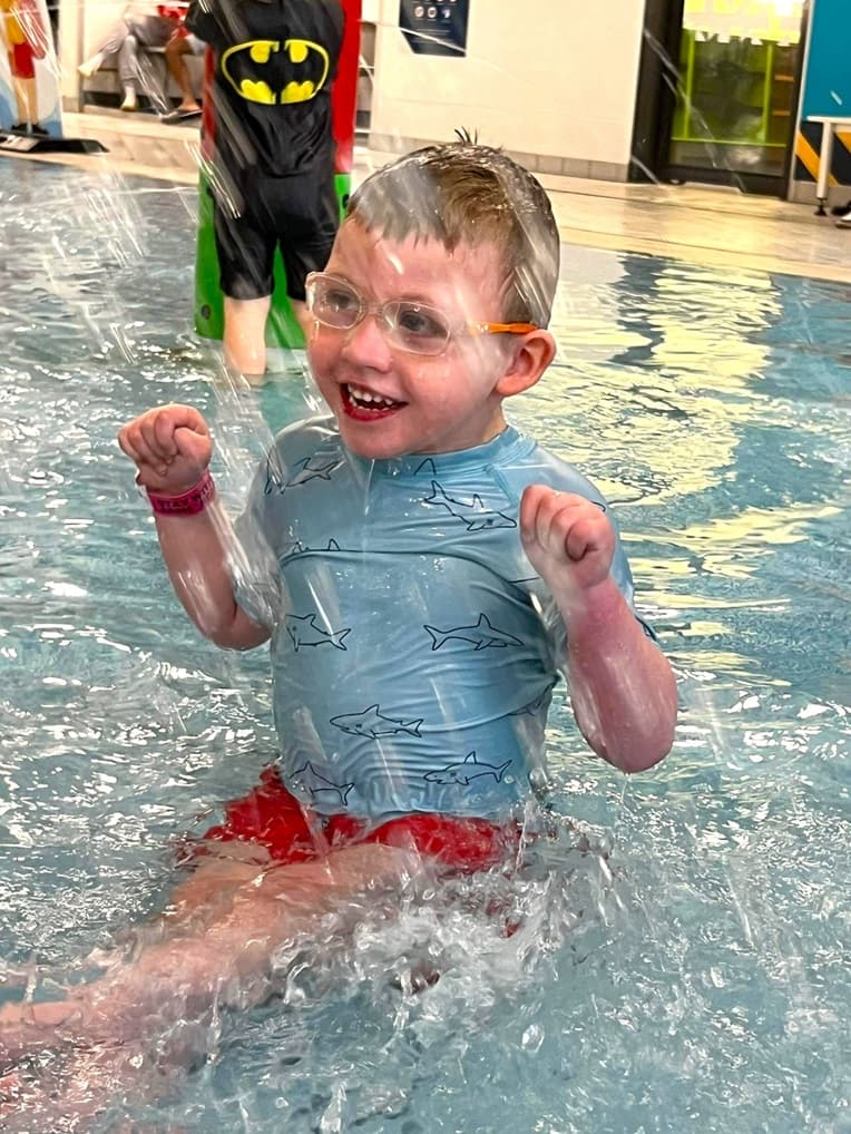 A smiling child wearing goggles splashes in a shallow indoor pool.