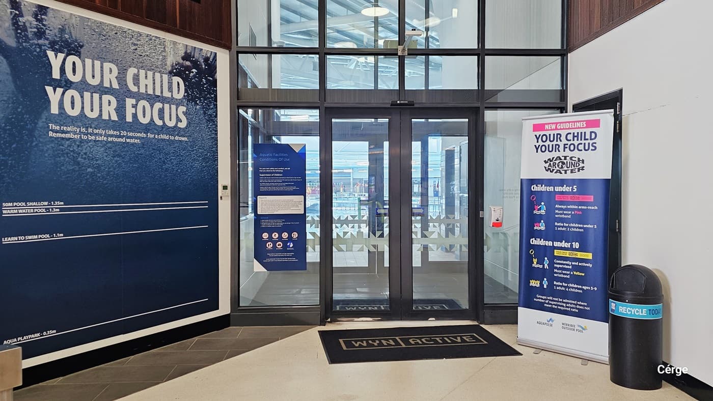 The Aquatic Centre entrance features two sets of automatic glass doors adorned with snowflake prints. A blue poster is affixed to the left section of the glass. A black doormat with "WYN | Active" is placed at the entrance. On the left, a large blue poster covers a significant portion of the wall. To the right, a banner displays child safety instructions, accompanied by a black rubbish bin. This view offers a glimpse of the indoor pool.