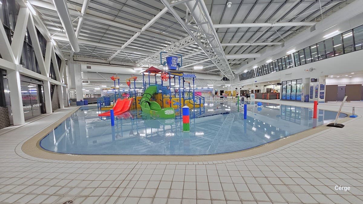 Another view of the shallow beach entry pool with two enclosed tube slides. The pool is equipped with interactive water cannons and a blue tipping bucket positioned overhead. The prominent white ceiling shows white pipes and tubes. There's a freestanding warning sign on the right. The floor is tiled in white. On the left, a glass wall features a large steel "W" design suspended from the ceiling. To the right, banners are on display, providing instructions and directions.