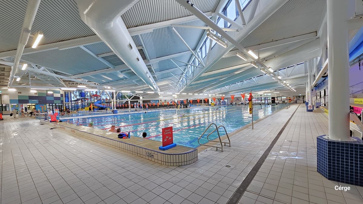 An alternate perspective of the heated pool reveals a stainless steel entrance ramp, accompanied by a freestanding orange safety banner positioned before the entrance. On the far left, a segment of the colossal winding water slide and the vibrant ceiling are visible. Orange triangular banners hang across the pool. On the right, there's a path leading to the opposite side of the pool. The floor and ceiling are both white, with large pipes and visible metal beams supporting the roof.
