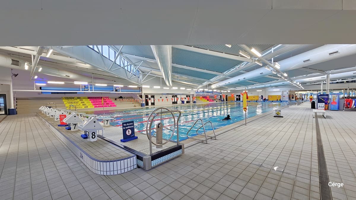 Another view of the heated pool displays a stainless steel entrance ramp and a platform featuring a stainless steel steering wheel, a freestanding safety sign, 8 stairs, and diving boards at the center. On the far left, there's a tiered platform with pink and yellow accents. To the right, additional safety signs and banners are visible, along with a chair placed in the middle. The floor and ceiling are both white, with large pipes and visible metal beams supporting the corrugated design roof.