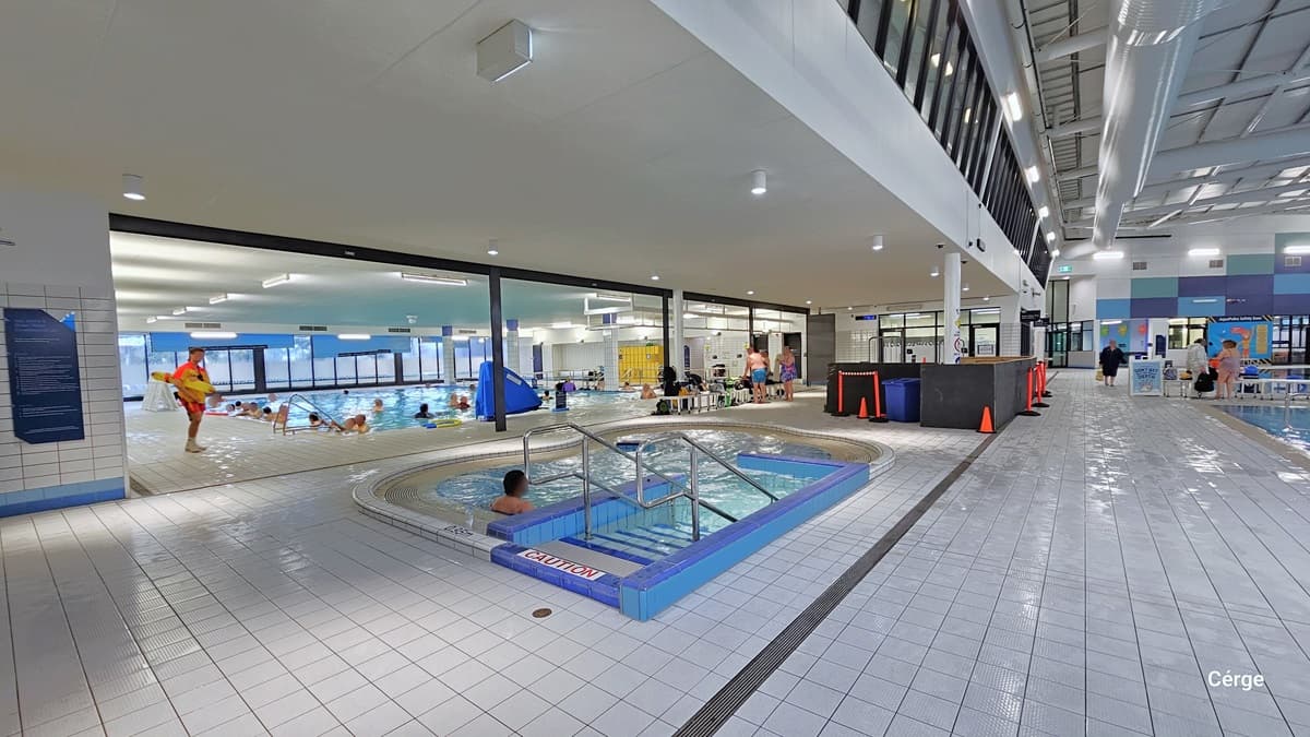 Here is a small spa pool in the middle, equipped with stairs and metal handrails. On the left, there is a larger pool, while on the right is another swimming pool. Adjacent to the spa pool is an area under construction, enclosed by black plywood barricades, orange cones, and rope bollards. The floor is tiled in white, and the ceiling is entirely white, with large pipes and visible metal beams supporting the roof. A few individuals can be seen swimming and moving about.
