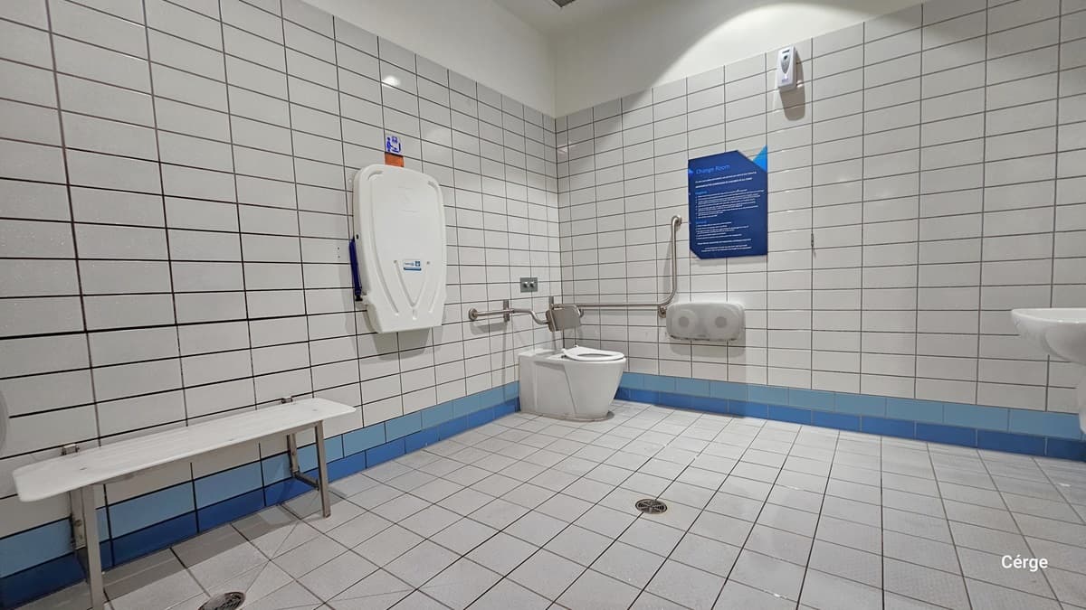 Inside the bathroom, there is a white bench on the left, a folding change table, and an ambulant toilet equipped with grab bars. In the middle, there's a tissue dispenser and an informational poster. On the right side, a wash basin is provided. The entire bathroom is tiled in white, including the wall with a blue accent, and the ceiling is painted white.