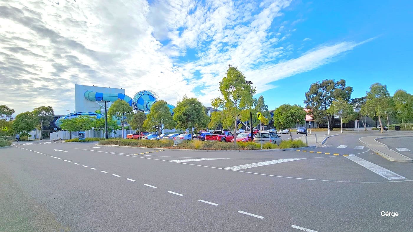 The AquaPulse building is visible, with its prominent spiraling water slide featuring shades of blue and green. The view encompasses both sides of the main road, one side revealing a pedestrian crossing. On the right, there's a turning road leading to the open car park area. The car park hosts a variety of cars in different colors and models. Medium-sized trees surround the parking area.