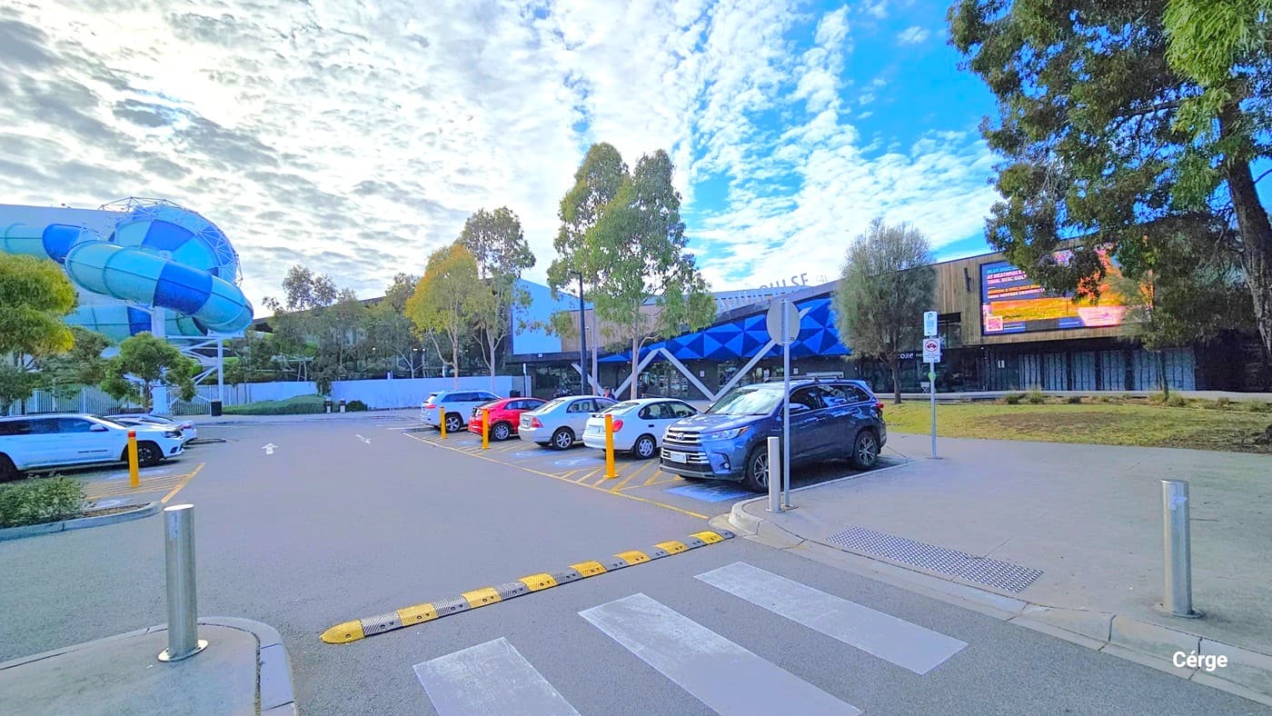 Seven marked accessible parking spaces are near the entrance, distinguished by yellow bollards and wheelchair-accessible signs. Seven cars of different models and colors are present. A marked pedestrian lane with white road markings and bollards borders the area. Truncated domes are placed for safety. The building's entrance is visible, with a large blue and green water slide on the left.