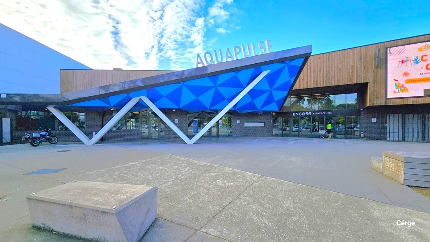 AquaPulse's main entrance has a distinctive design with the letter "W" seemingly supporting a triangular ceiling in shades of blue and green. The name "AquaPulse" is prominently displayed on the rooftop. On the right, a large digital screen is visible. In front, there's a spacious area with a raised concrete structure in the center. The building's exterior features a combination of grey and brown wooden design. A motorcycle is parked to the left.