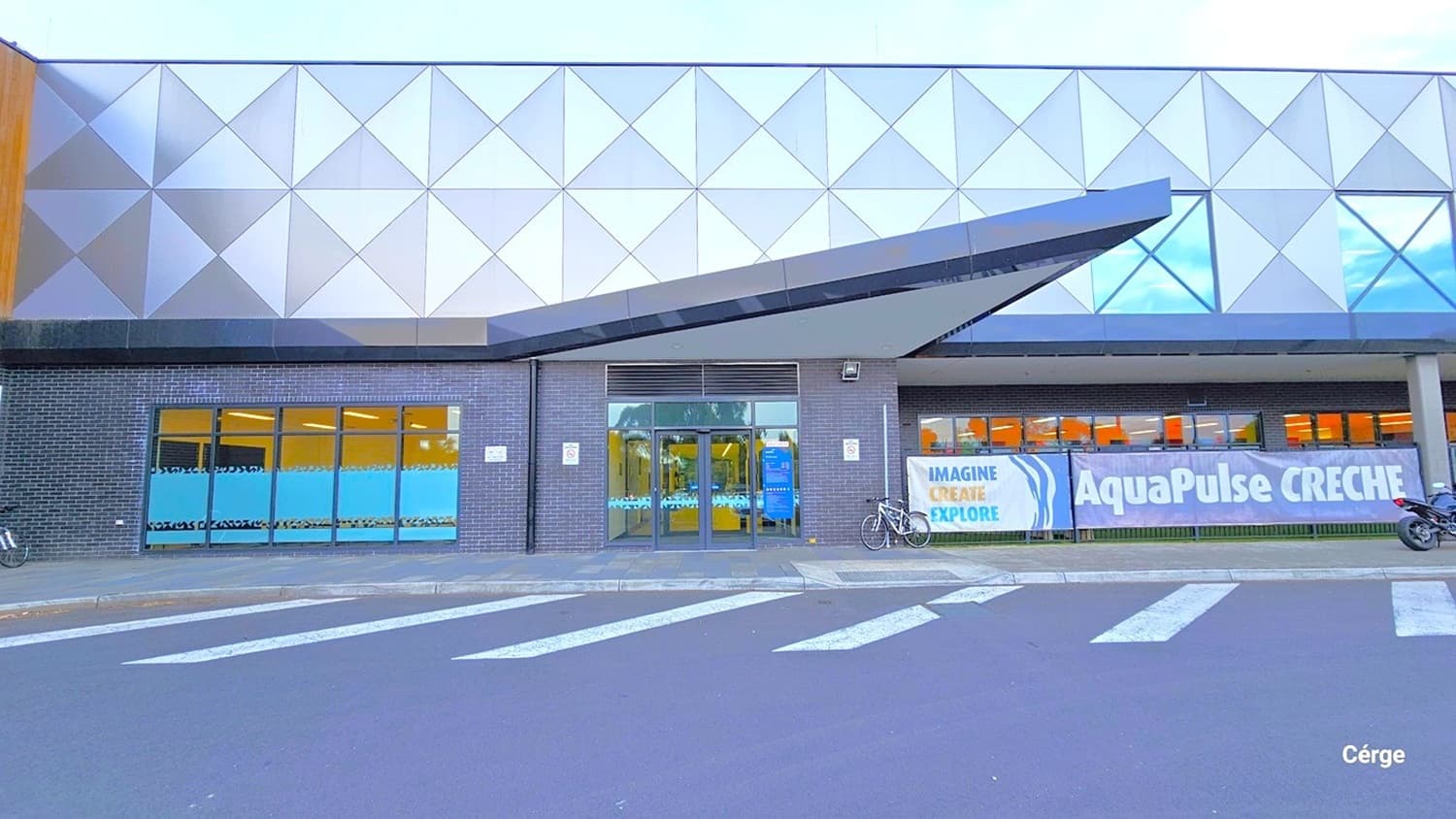 The step-free entrance to AquaPulse is situated on the left side of the main entrance, marked by a concrete ramp and pedestrian road markings. From this vantage point, you can observe the distinctive design of the building's exterior with wall prints. Adjacent to the automatic door, there's a lengthy banner reading "AquaPulse Creche," along with another blue banner displaying instructions. Two bicycles, including a motorcycle, are positioned near the entrance.