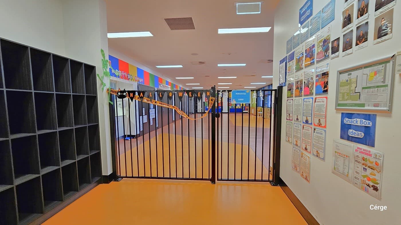 This area comprises an entrance and exit secured by a black-painted steel gate. To the left, there are black 4-tier open cabinets, and to the right, a white wall adorned with posters displaying instructions, reminders, tips, and helpful information. The floor is tiled in yellow. From this viewpoint, you can observe the interior of the Creche, where colorful paper decorations adorn the walls.