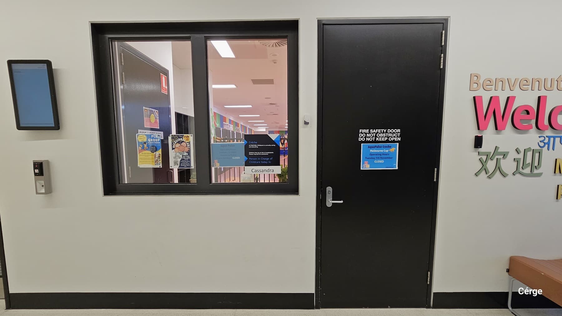 The Creche at AquaPulse. In front, there is the entrance to the creche, featuring a brown door with a stainless steel handle. The door bears printed instructions, reading "Fire Safety Door, Do not obstruct. Do not Keep Open." Another blue poster with instructions is affixed to the door. To the left, there's a sliding window with additional posters, an emergency light, and a controller. A brown cushioned chair is positioned near the door, and the wall displays inscriptions in various languages.