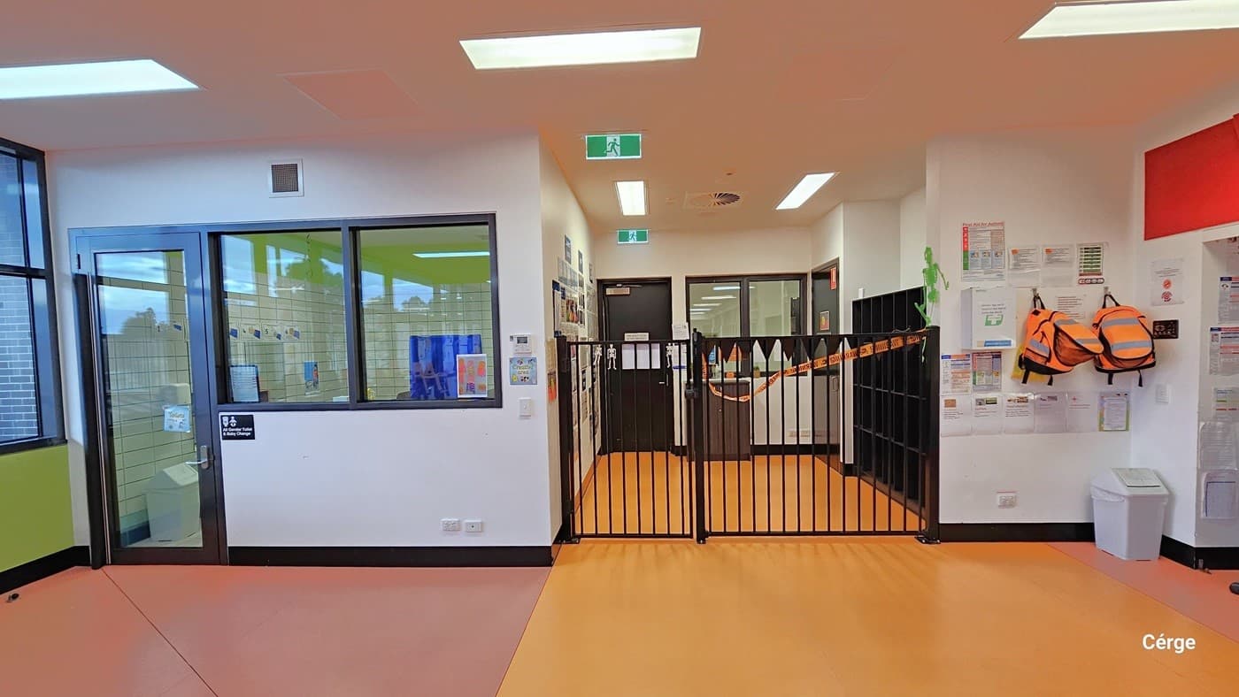 Next to the Creche entrance, there is the accessible all-gender toilet on the left, enclosed by a large see-through window and door. In the center is a low brown steel gate adorned with Halloween banners, and another door is visible behind it. On the left, posters are affixed to the white wall, while two orange backpacks are hanging, with a white rubbish bin below. The white ceiling features two green fire exit signs, and the floor consists of rubber tiles in pink and yellow.