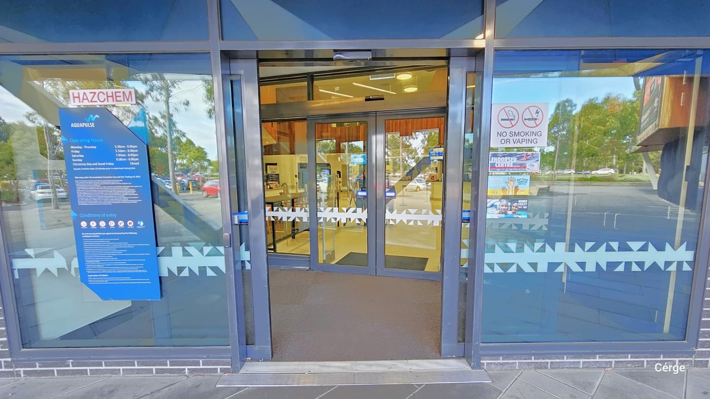 AquaPulse entrance is through two sets of automatic glass sliding doors fixed within silver railings. Stickers and banners on both doors display reminders and instructions, including a "No smoking or vaping" sign. The glass doors feature snowflake design markings. Within sight is a section of the interior cafe. whose interior is well-illuminated.