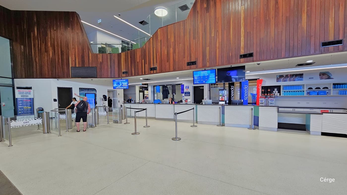 Upon entry, the reception area is straight ahead, featuring queue barriers, digital information screens, and merchandise for sale. The entrance includes a turnstile where two individuals are seen entering. The space is well-lit with a white ceiling and floor tiles. The wall showcases an intricate brown wooden design. You can also see the second-floor area from this perspective.