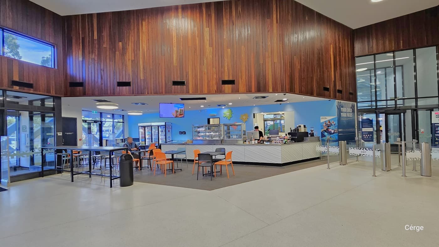 To your left is a café with tables, black and orange chairs, and a long high table with stools. A black rubbish bin sits at the table's end. The counter holds food display racks and a drinks fridge. A cashier stands by the POS computer. To the right, the entrance/exit area is enclosed with fiberglass featuring a snowflake design. The space is well-illuminated, with a white ceiling and floor tiles. The wall features an intricate brown wooden design, and you can see the second-floor area.