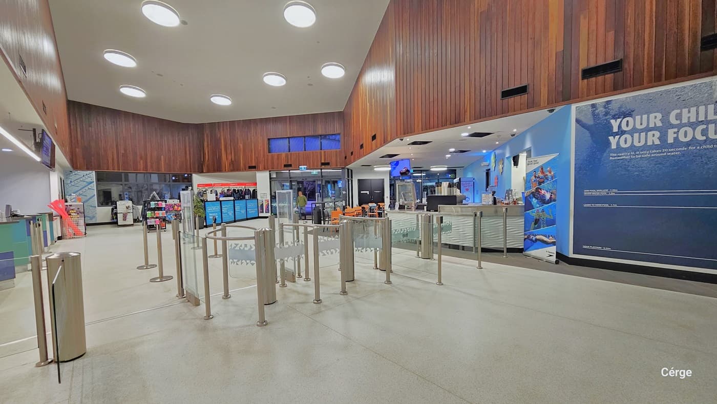 Upon entry, the reception area features queue barriers, digital information screens, and merchandise for sale. The entrance has a turnstile with two people entering. Well-illuminated space with a white ceiling, floor tiles, and an intricately designed brown wooden finish on the wall. Further ahead, a retail area offers swimming costumes, goggles, and swim accessories.