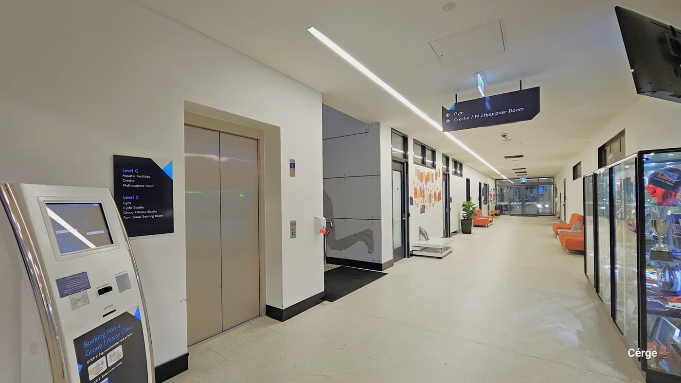 The lobby provides access to the Creche/Multipurpose Room. On the eft, there's a computer machine for booking fitness classes, alongside a cream-colored elevator with an information poster. Next to the elevator is a staircase. The left walls are covered with colorful papers, and a banner on the ceiling indicates directions. To the right, a retail shop section showcases trophies in a glass cabinet. The area is well-lit, with white flooring, ceiling, and walls.