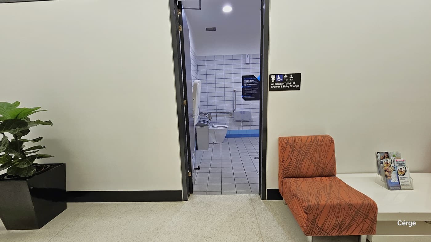 Entrance to the All-Gender Toilet. On the left, there's an ornamental plant in a large black plant box. To the right, there is a cushioned brown chair, a white table, and a few magazines on top. The wall before entering the area has a sign indicating it's an all-gender toilet with the inscription "All Gender Toilet Shower and Baby Change" affixed to the white wall. From this view, you can see the interior of the toilet area, with white tiled floors and walls.