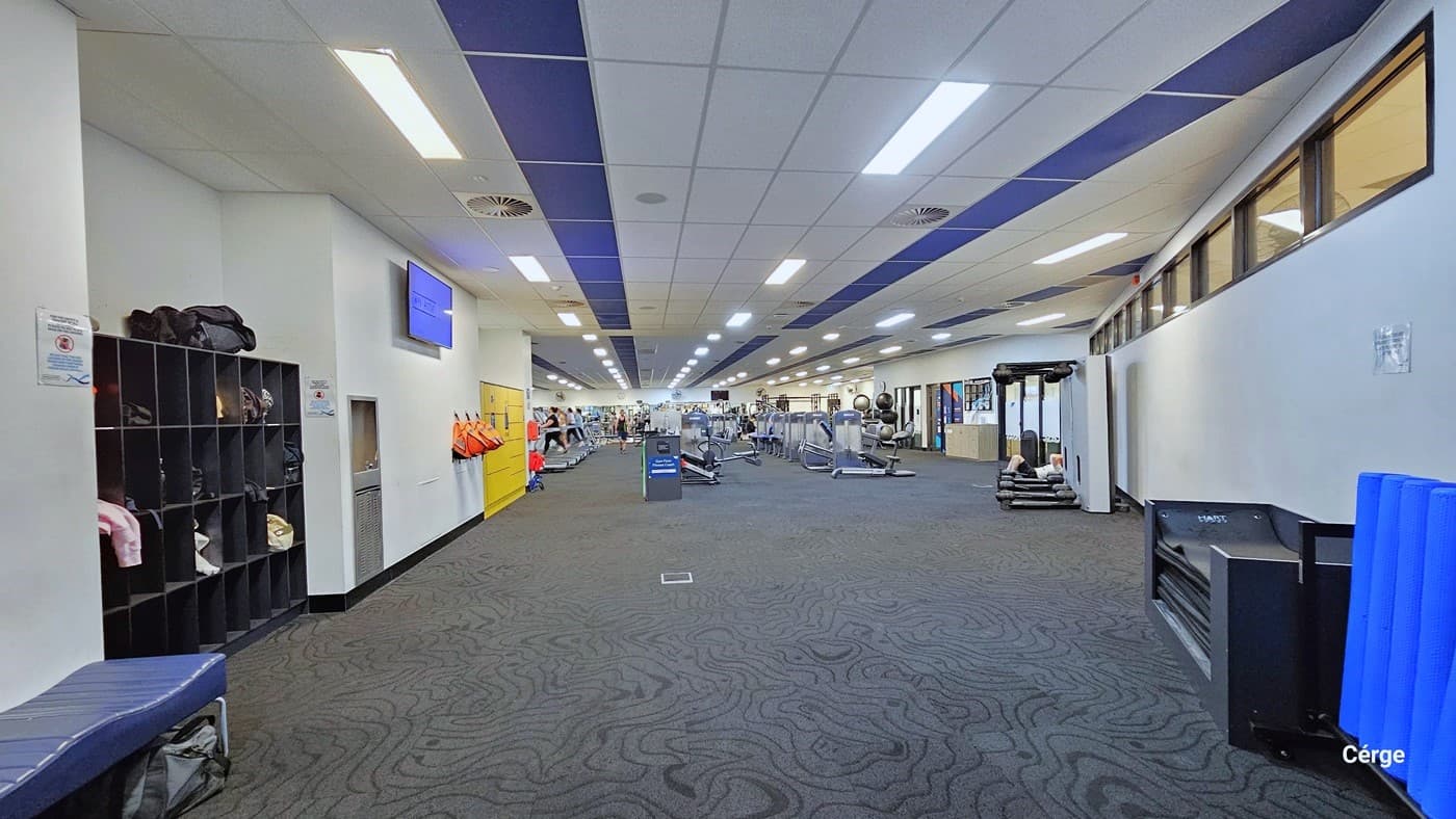 In the gym area, you'll find storage units, yellow lockers, and a blue cushioned bench on the left. A flat-screen TV is wall-mounted. On the right, there's another bench with blue pillows. Further ahead are rows of exercise machines. The area has grey carpets, white ceiling lights, and centralized air conditioning.