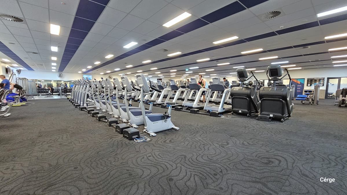 AquaPulse Gymnasium. This expansive area is filled with rows of cardio machines and various fitness equipment, including treadmills, rowing machines, and stair climbers neatly arranged in rows. More exercise equipment is visible to the left. The area is carpeted in grey, and the white ceiling is fitted with centralized air conditioning and fluorescent lights. There are a few people who are using treadmills.