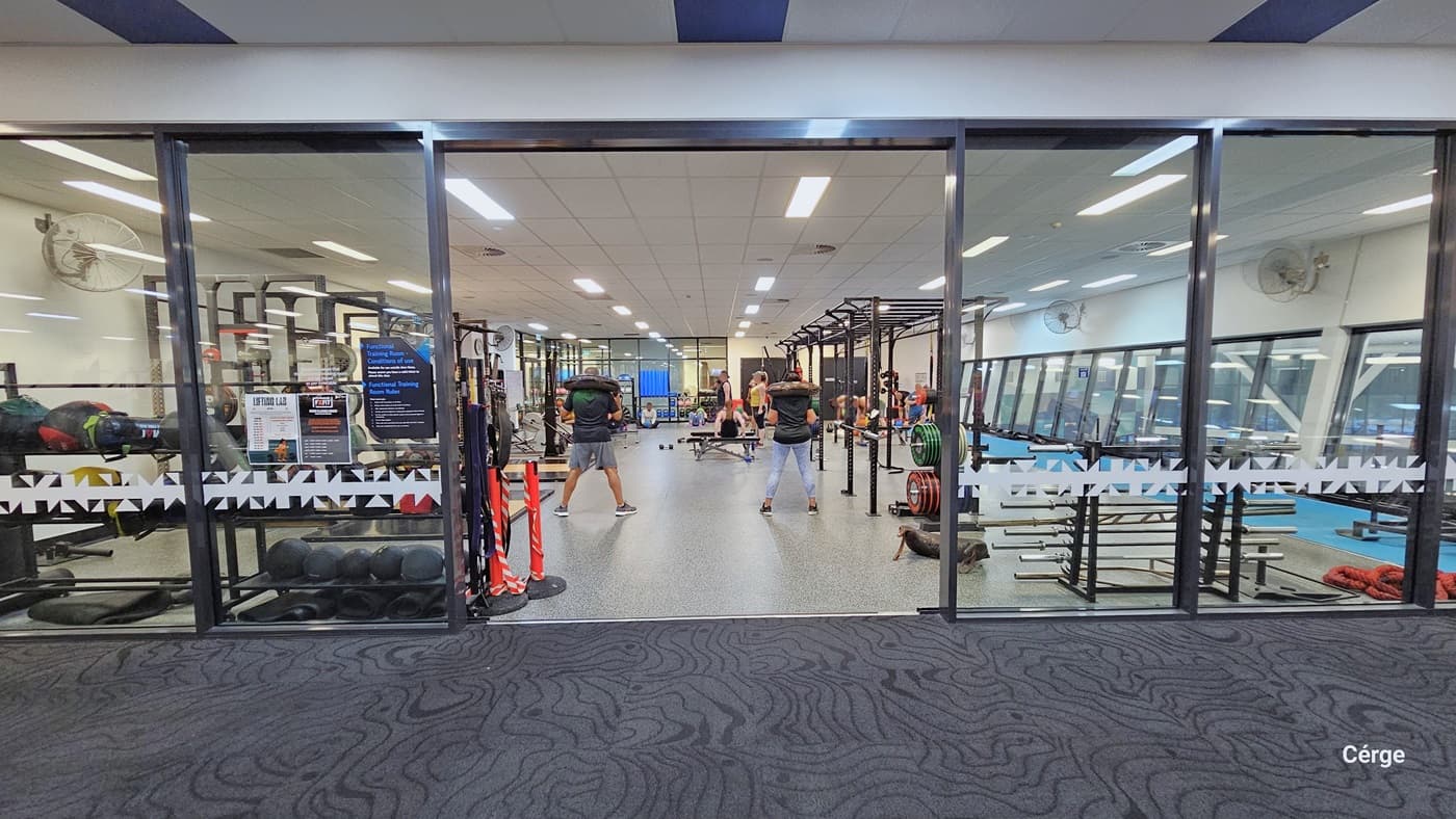 This view displays the opposite side of the gym, dedicated to muscle strength training. Various strength machines are neatly arranged on the right, while more are on the left. Orange bollards are positioned at the door entrance, and a fan is mounted on the left wall. The floor area combines carpet and timber. The area features grey carpeting, and the white ceiling is equipped with centralized air conditioning and fluorescent lights. A few people are exercising.