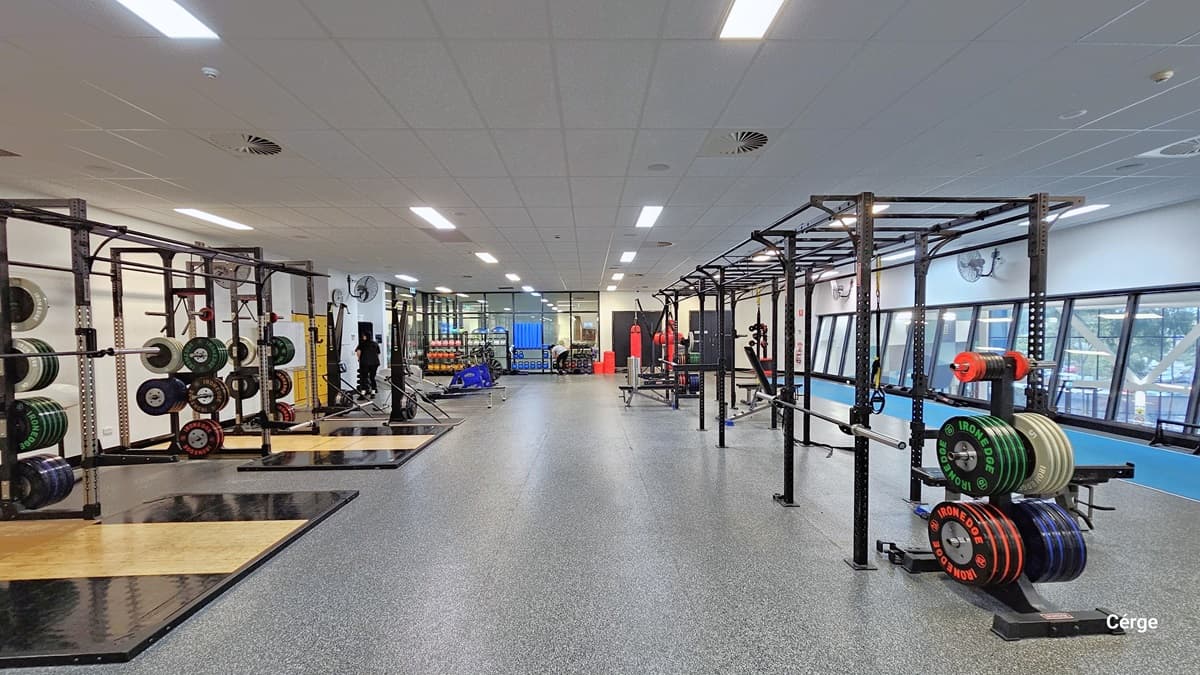 This view reveals a spacious gym area with Smith machines on both sides. Weights are neatly arranged. Further ahead, more strength machines are visible. The area has grey rubber tiles, and the white ceiling is equipped with centralized air conditioning and fluorescent lights. Yellow lockers can be seen on the far left.