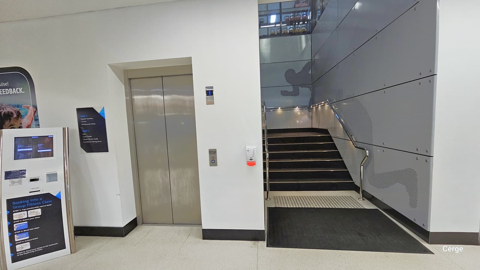 Entry to Health Club Level 1. To the left, there is a lift with a wall-mounted sign indicating direction. Two banners are placed before the lift entrance. On the right, there's a staircase leading to Level 1, featuring a black non-slip carpet, white truncated domes positioned before the stairs, stainless handrails, and black carpet. The wall is painted in grey with a faint shadow design of a running man.