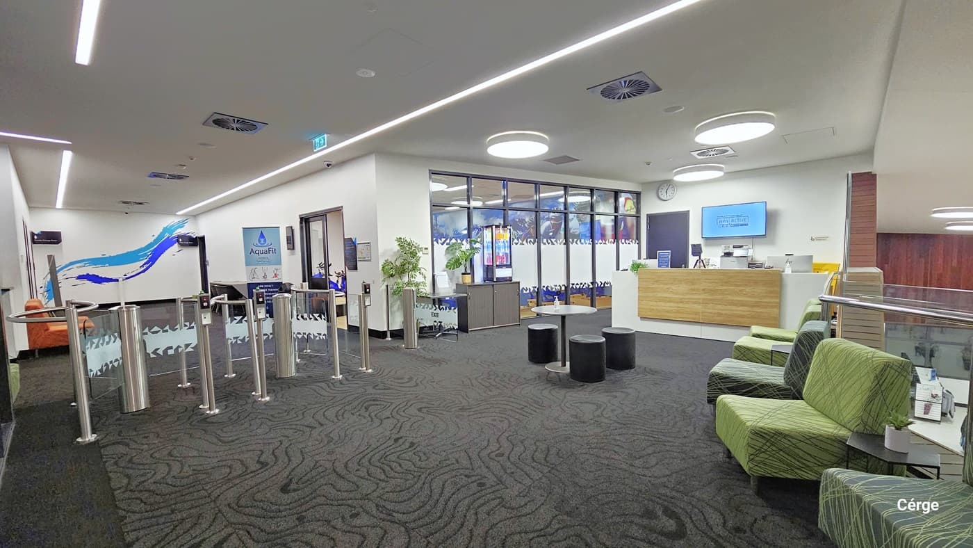 Entry to Health Club. Turnstiles are installed at the entrance. To the right is the cycle studio. There is a reception desk on the right. A round table and round cushioned stool are in the center, with cushioned green chairs lined up on the sides. The area has grey carpeted floor, an all-white ceiling, and white walls with minimal blueprints. The ceiling features designed lights and a few centralized air conditioning systems.