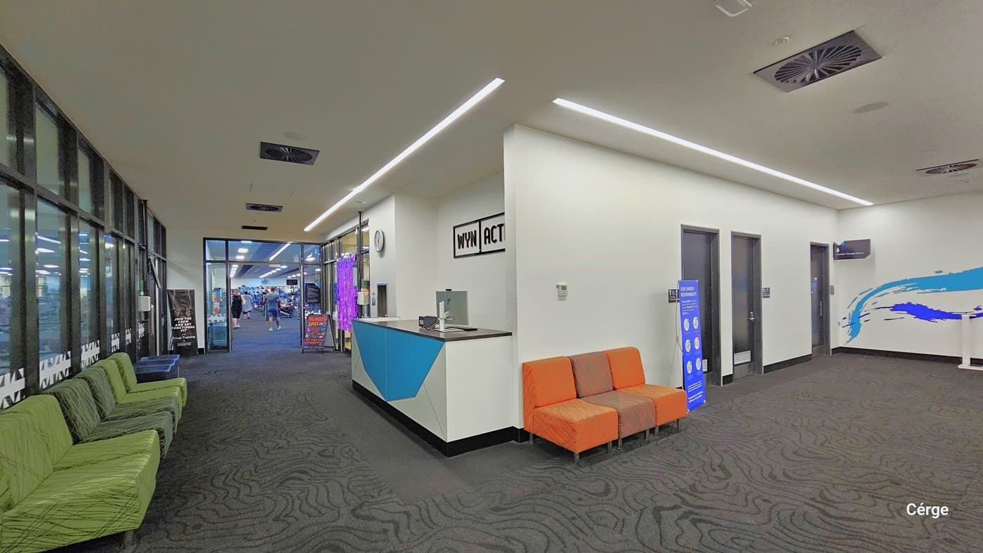 To the right, there are black doors, two of which lead to all-gender toilets. In the middle, there are three cushioned orange chairs and a reception desk. On the left, a few green cushioned chairs are lined up against the glass wall. The area has grey carpeted floor, an all-white ceiling, and white walls with minimal blue prints. The ceiling features designed lights and a few centralized air conditioning systems.