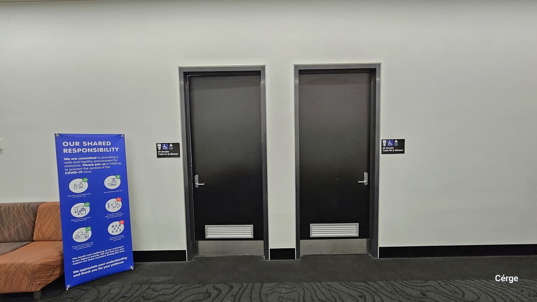 The bathrooms. Two black doors are marked as all-gender toilets, accessible to disabled individuals, and equipped with a shower. On the left, several orange cushioned chairs are arranged against the glass wall. Next to the chairs, there's a blue free standing banner displaying reminders. The area features a grey carpeted floor, an all-white ceiling, and white walls.